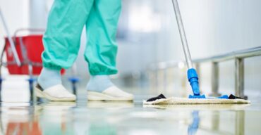 Profissional realiza higiene e limpeza hospitalar com mop em corredor de unidade de saúde.