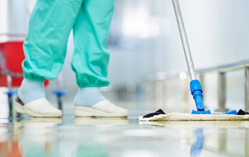 Profissional realiza higiene e limpeza hospitalar com mop em corredor de unidade de saúde.