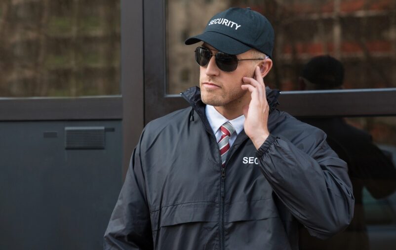 Vigilante patrimonial atento com uniforme preto em frente a prédio.