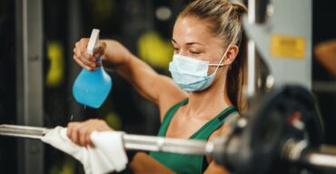 Mulher faz a higienização de academia usando borrifador e pano em barra de musculação.