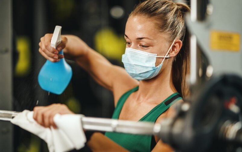 Mulher faz a higienização de academia usando borrifador e pano em barra de musculação.